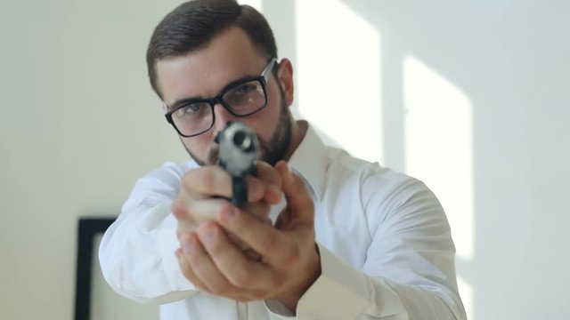 closeup portrait of athletic businessman with beard and eyeglasses in white shirt holding black gun pointing at camera professional private job security guarantee man shotter safety dangerous guy