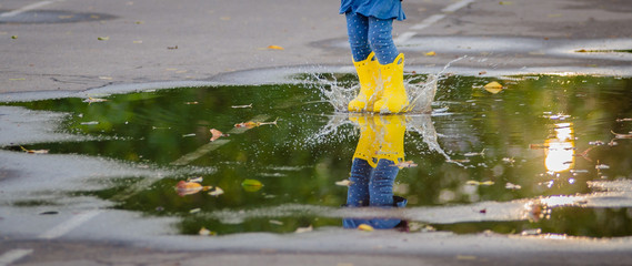 Happy funny child with multicolored umbrella jumping puddles in rubber boots and laughing