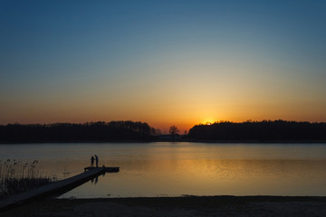 Pier with silhouettes of two fishermen on the background of a lake in which the evening sky and sunset are reflected.