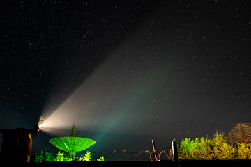 Beam of an old searchlight and line of barbed wire against the background of the big antenna of the radio telescope illuminated by green light under the night star sky.