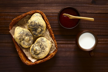 Homemade poppy seed bread rolls in basket with cup of milk and strawberry jam on the side, photographed overhead on dark wood with natural light