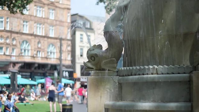 Establishing shot of the Shakespeare Memorial Fountain in Leicester Square, London, with fish sculptures and water sprays and out of focus people in the background.