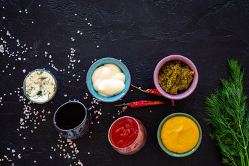 Different dip sauce in bowls near chili pepper and greenery. Ketchup, mayonnaise, mustard, soy sauce, barbecue sauce, pesto, mustard, sour on black background top view copy space