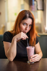 Young attractive red hair woman in a black t-shirt is drinking a milkshake in a cafe. Young girl drinking fruit smoothie in cafe