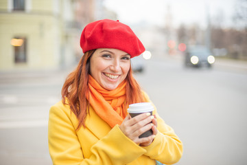 the red-haired young woman in a yellow coat and red beret is drinking coffee on the street to keep warm. A girl in bright outer clothes drinks hot tea with delight