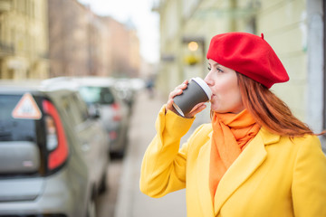 the red-haired young woman in a yellow coat and red beret is drinking coffee on the street to keep warm. A girl in bright outer clothes drinks hot tea with delight