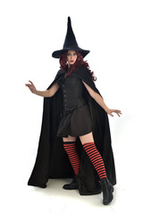 full length portrait of red haired girl wearing long black cloak, pointy hat and witch costume. standing pose, isolated on white studio background.