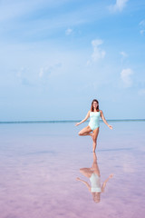 A beautiful woman in a mint body stands in a tree position in a pink salt lake. Redhead young woman doing yoga standing ankle-deep in water, pink salt lakes Bursol'