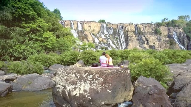 couple sits on rock among river rapids by waterfall