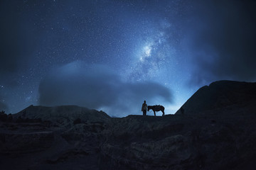 Fototapeta premium Horsemen standing under million stars in sky with his horse near smoking hot volcano Mount bromo at night, Indonesia