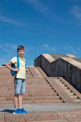 a boy with a school bag stands tall against a blue sky, to the right of the stairs.