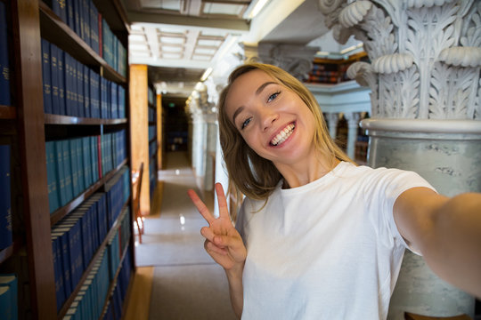 Excited young girl standing in traditional old library at bookshelves, smiling and laughing student making selfie on phone camera, having fun. Higher education and Student life concept.