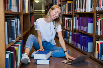 Young girl sitting on the floor in traditional library at bookshelves, reading books. Smiling and laughing student working with laptop, studying. Higher education.  Student life