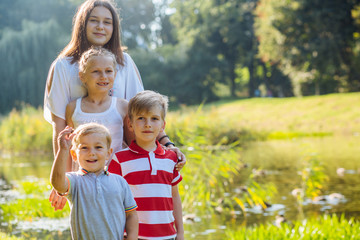 Four children of different age - teenager girl, school girl, elementary age boy and preschooler boy standing and hugging by a forest lake looking at camera smiling. Copy space