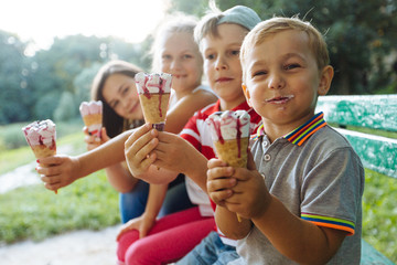 Group of four happy children eating ice cream together outdoor. Photo of happy blond girls with two handsome boys sitting on the bench and smiling at camera. Sun glare effect - Powered by Adobe