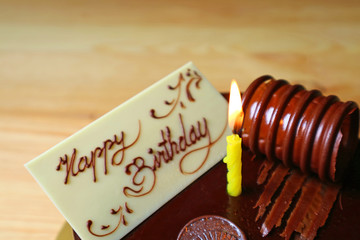 White Chocolate Greeting Card with a Lighting Yellow Candle on Chocolate Mousse Birthday Cake on the Wooden Table 