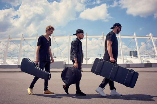 side view of rock music band with musical instruments in cases walking on street with cloudy sky on background