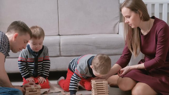 Family mom, dad and two twin brothers play together building out of wooden blocks on the floor.