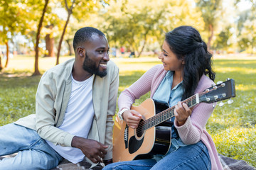 smiling african american girlfriend playing acoustic guitar for boyfriend in park