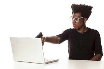Angry and nervous young african-american guy using laptop computer, aiming with a gun working project at desk on white background