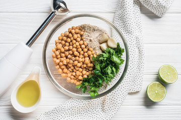 Flat lay with chickpeas, parsley and spices ingredients for hummus in bowl, blender and lime pieces on wooden surface