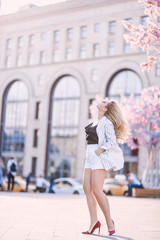 Naklejka premium Portrait Beautiful young blonde girl in sunglasses with puffy lips and sexy feminine body posing on the street with pink flowers on the background on sunny day 