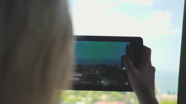 A young girl with blonde using a tablet takes pictures and videos of the landscape outside the window.