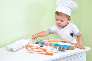 A cute two year old boy dressed as a chef makes cookies from Ingredients that are on his desk.