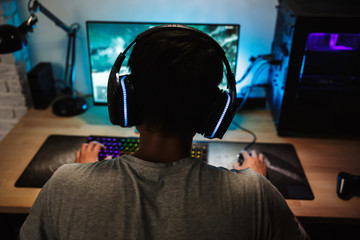 Back view of teenage gamer boy playing video games online on computer in dark room, wearing headphones with microphone and using backlit colorful keyboard © Drobot Dean