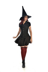 full length portrait of red haired girl wearing black witch costume and pointy hat.  standing pose, isolated on white studio background.