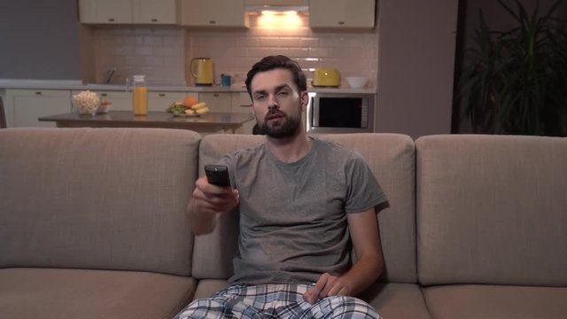 Hansdsome man sits on sofa and watches TV. He uses remote control several times. Man pushes buttons on it. Finaly he stopped doing that and strated to smile.