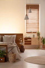 Wooden cupboard next to bed in bedroom interior with lamps and blinds on window. Real photo