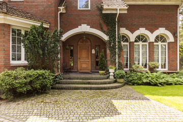 Cobbled path and steps leading to a stylish entryway with ornamented wooden door and side windows in a red brick English style mansion.