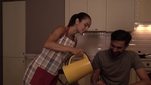 Careful girl wears pron and holds yellow kettle. She pours hot water in cup of her husband first and then into cup of herself. He is happy and smiles. And girl as well.