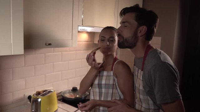Girl stands with guy. She holds toast with nutella on it. THen girl is giving it to man. He bites piece and starts to chew. She is looking at him and smiling. Then girl bites a piece from toast too.