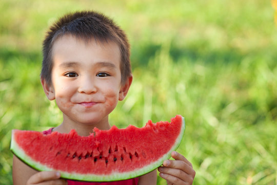 Happy Smiling Asian Child Eating Watermelon In The Garden. Kids Eat Fruit Outdoors. Healthy Snack For Children. Little Boy Playing In The Garden Biting A Slice Of Watermelon.