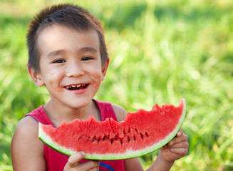 Happy smiling asian child eating watermelon in the garden. Kids eat fruit outdoors. Healthy snack for children. Little boy playing in the garden biting a slice of watermelon.