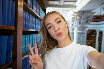 Excited young girl standing in traditional old library at bookshelves, smiling and laughing student making selfie on phone camera, having fun. Higher education and Student life concept.