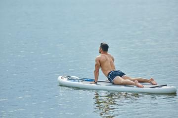 Fit man doing stretching exercises laying on sup board, swimming on city lake.