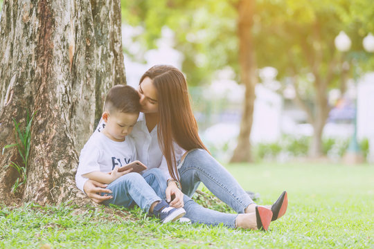 Little boy concentrate at smartphone with his mom kiss and sit together on green yard under giant tree.
