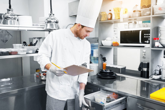 cooking, profession and people concept - male chef cook with clipboard doing inventory and looking to fridge at restaurant kitchen