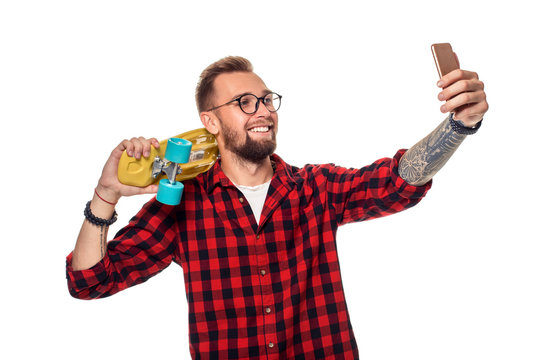 Young man holding the skateboard on shoulder raising his phone takes a selfie on a white background.
