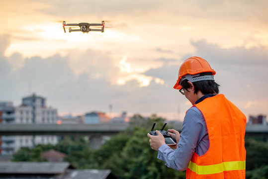 Young Asian engineer flying drone over construction site during sunset. Using unmanned aerial vehicle (UAV) for land and building site survey in civil engineering project.