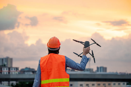 Young Asian engineer man holding drone at construction site during sunset. Using unmanned aerial vehicle (UAV) for land and building site survey in civil engineering project.