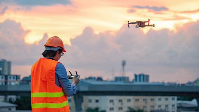 Young Asian engineer man flying drone over construction site during sunset. Using unmanned aerial vehicle (UAV) for land and building site survey in civil engineering project.