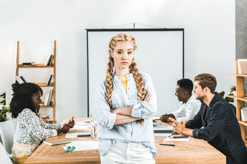 portrait of upset businesswoman with arms crossed looking at camera and colleagues working behind...