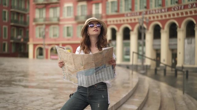 Attractive young woman tourist with long brown hair wearing a hat and purple sunglasses is standing in street of Nice, France with a map and looking around. Handheld slow motion medium shot