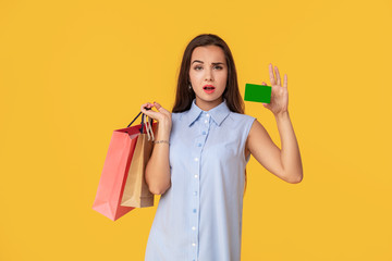Trendy woman 20s in dress with long brown hair smiling while holding different shopping packages and card in hands isolated over yellow background