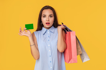 Fototapeta na wymiar Charming pretty attractive sexy girl showing credit card and holding many colorful packages in hand, it is comfortable to use bank card for shopping, standing over yellow background