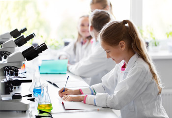 education, science and children concept - kids studying chemistry at school laboratory and writing to workbooks